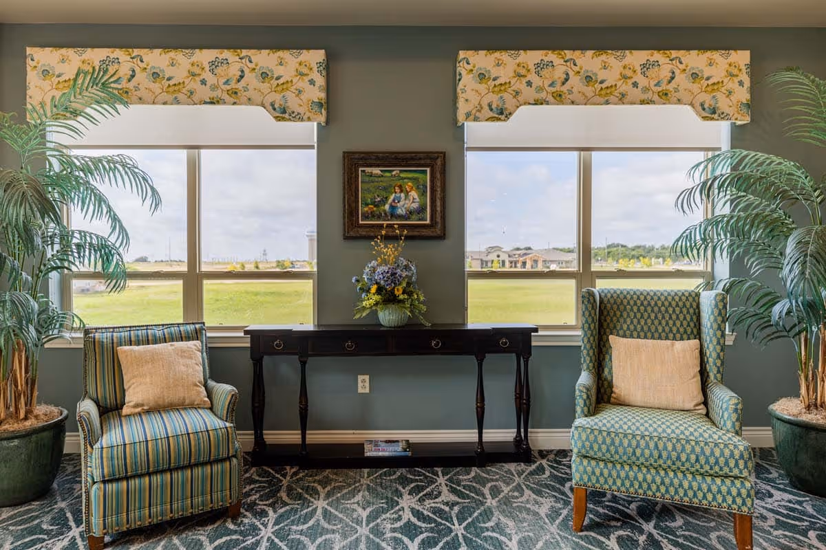 A cozy sitting area in a living room with two upholstered armchairs, one striped and one patterned, each with a beige cushion. Between the chairs is a dark wooden console table with a floral arrangement on top. Two large windows with floral valances provide a view of a grassy outdoor area. Two large potted plants flank the seating area, and a framed painting hangs on the wall between the windows.