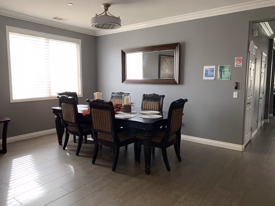 A dining room with a dark wooden table and six matching chairs with striped upholstery. The table is set with placemats and decorative candles. A large window with blinds allows natural light to fill the room. A rectangular mirror with a dark wooden frame hangs on the gray wall. The floor is covered with light-colored wood-like tiles. There are some signs and a no smoking sign on the wall near a hallway with multiple doors.