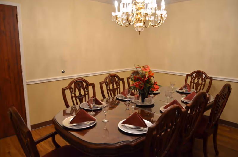A formal dining room with a polished wooden table set for eight people. Each place setting includes a white plate, a folded maroon napkin, and a wine glass. A floral centerpiece with red and orange flowers is placed in the middle of the table. The room has beige walls with white trim and a wooden door on the left. A chandelier with multiple lights hangs above the table.
