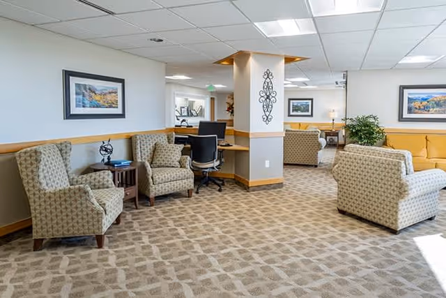 A spacious and well-lit living area in a senior living facility featuring patterned carpet, several upholstered armchairs, a desk with an office chair, framed landscape artwork on the walls, and decorative wall art on a column. The room has a neutral color palette with beige and light brown tones.