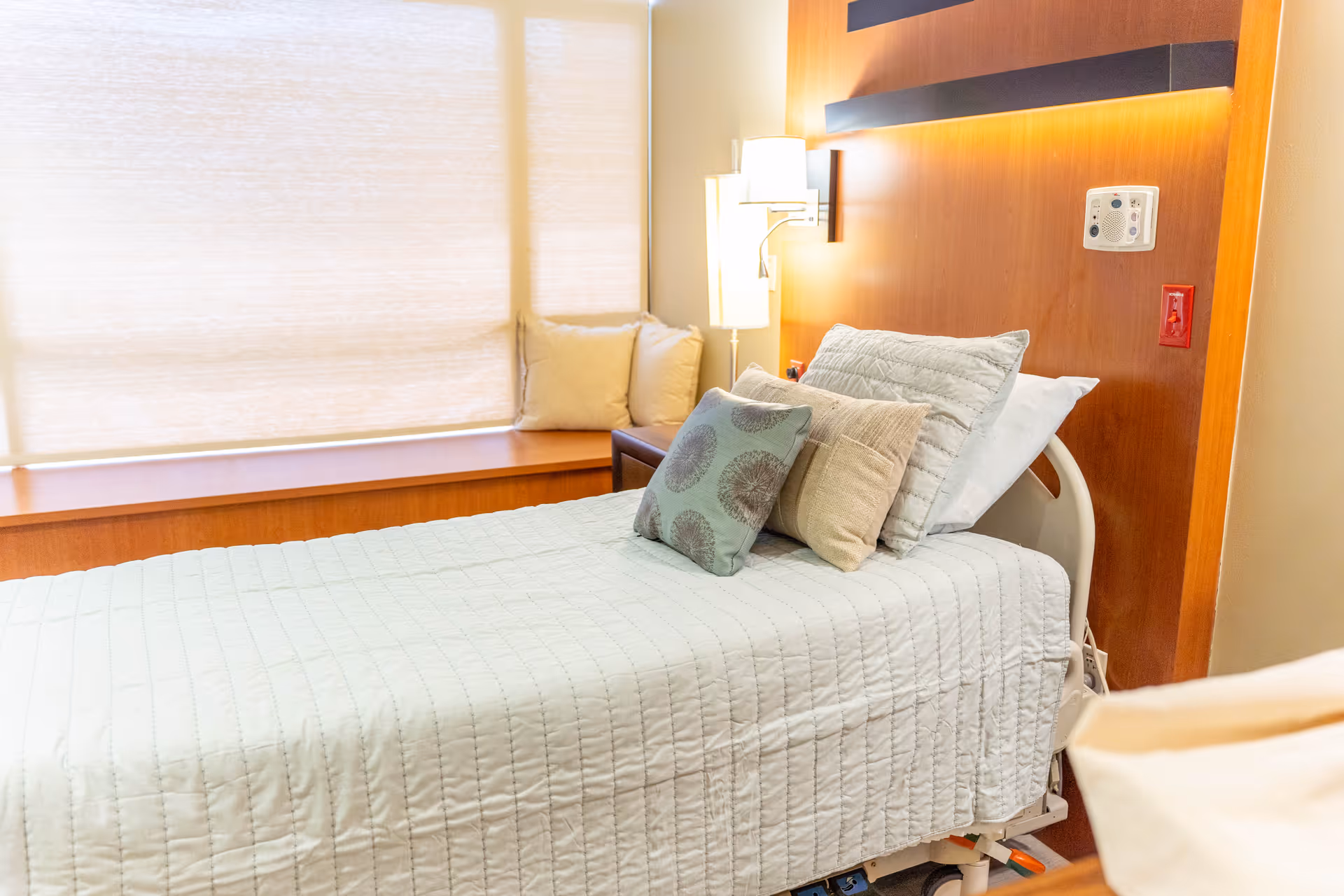 A neatly made hospital-style bed with multiple pillows in a softly lit room. The bed has a white quilted cover and is positioned next to a wooden headboard with built-in lighting and a call button panel. There is a window with closed blinds and a cushioned bench beneath it, along with a floor lamp and additional pillows in the corner.