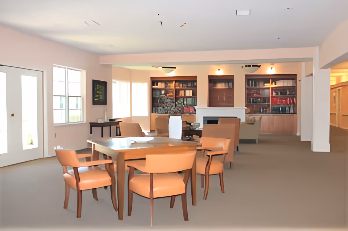 Bright communal dining and lounge area with a wooden table and upholstered chairs, bookshelves and seating near a fireplace.