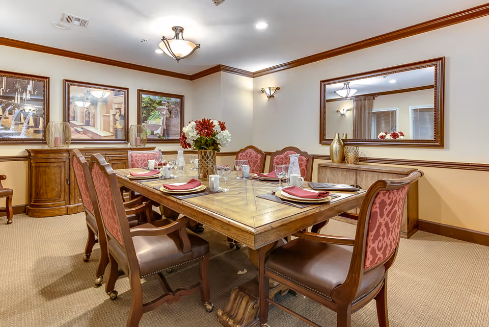 A formal dining room with a large wooden table set for six people. The table has red napkins, white plates, glassware, and a centerpiece with red and white flowers. The room features wooden trim, beige walls, carpeted floor, framed artwork on one wall, and a large mirror on another wall reflecting part of the room.