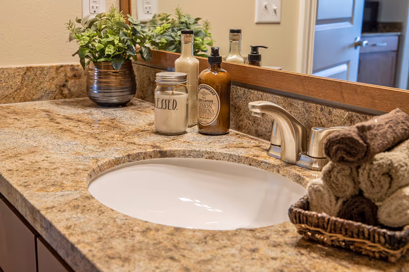 A bathroom countertop with a built-in sink, a silver faucet, a small potted plant, a candle jar labeled 'BLESSED.', a bottle of lemon verbena hand lotion, and a basket holding rolled brown and beige towels. A large mirror is mounted on the wall behind the countertop.