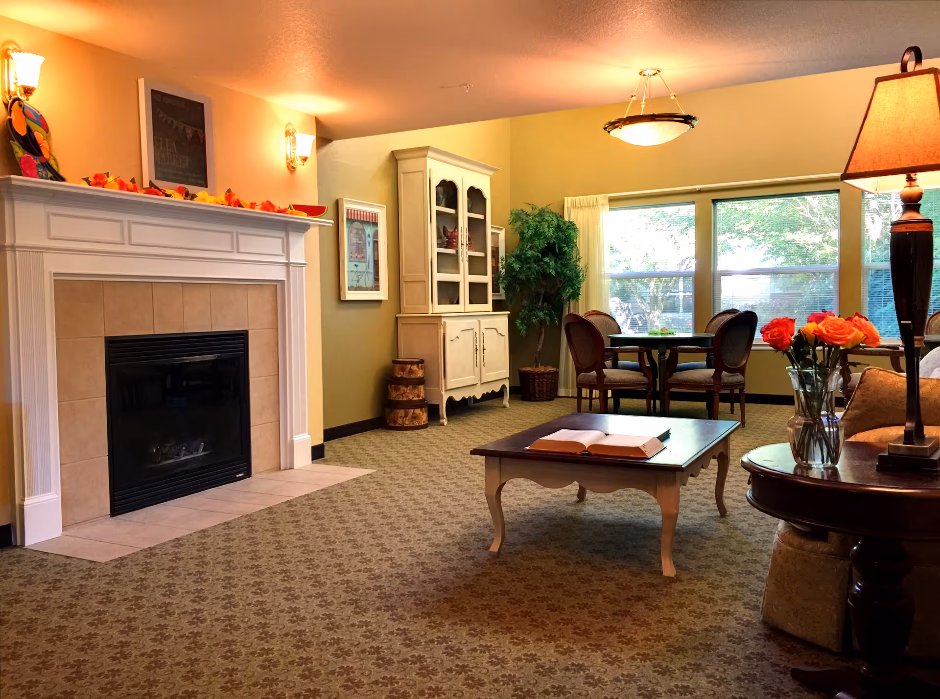 A cozy senior living room with a fireplace on the left, decorated with autumn leaves and wall sconces. There is a white cabinet against the back wall, a round dining table with four chairs near large windows, and a coffee table with an open book in the center. A vase with orange roses and a lamp are on a side table to the right.