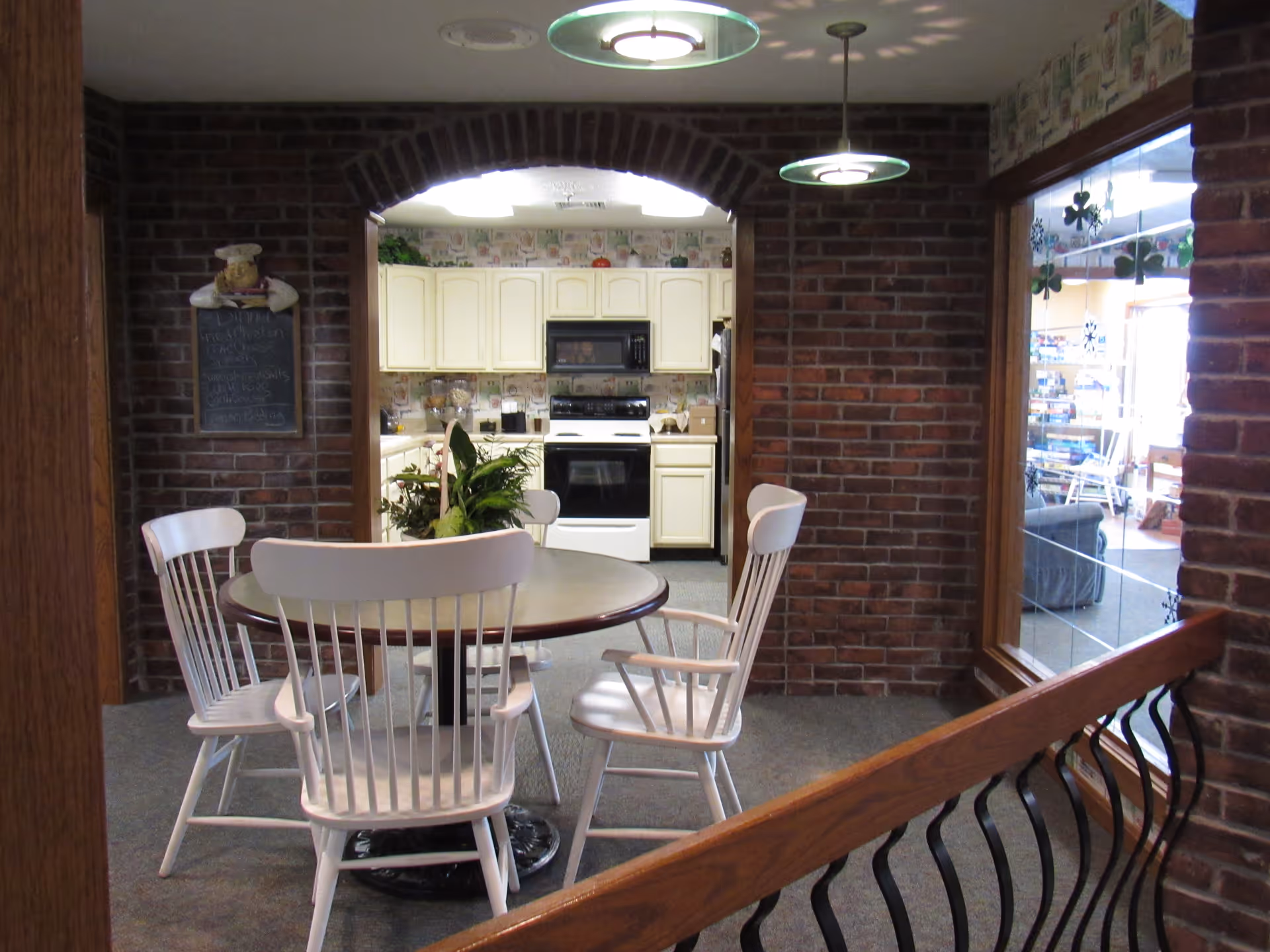Interior view of a dining area with a round table and four white wooden chairs. The dining area is adjacent to a kitchen with white cabinets, a stove, and a microwave. The walls are brick with an arched doorway leading to the kitchen. There is a window on the right side showing another room with seating and decorations.