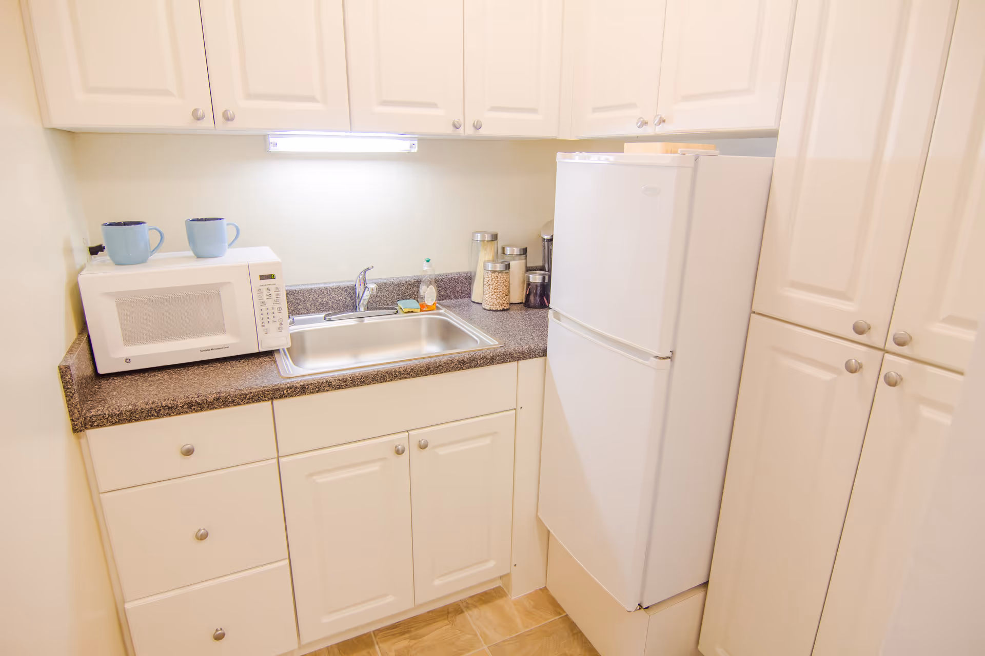 Small kitchen area with white cabinets, a white refrigerator, a microwave with two blue mugs on top, a stainless steel sink, and several jars containing food items on the countertop.