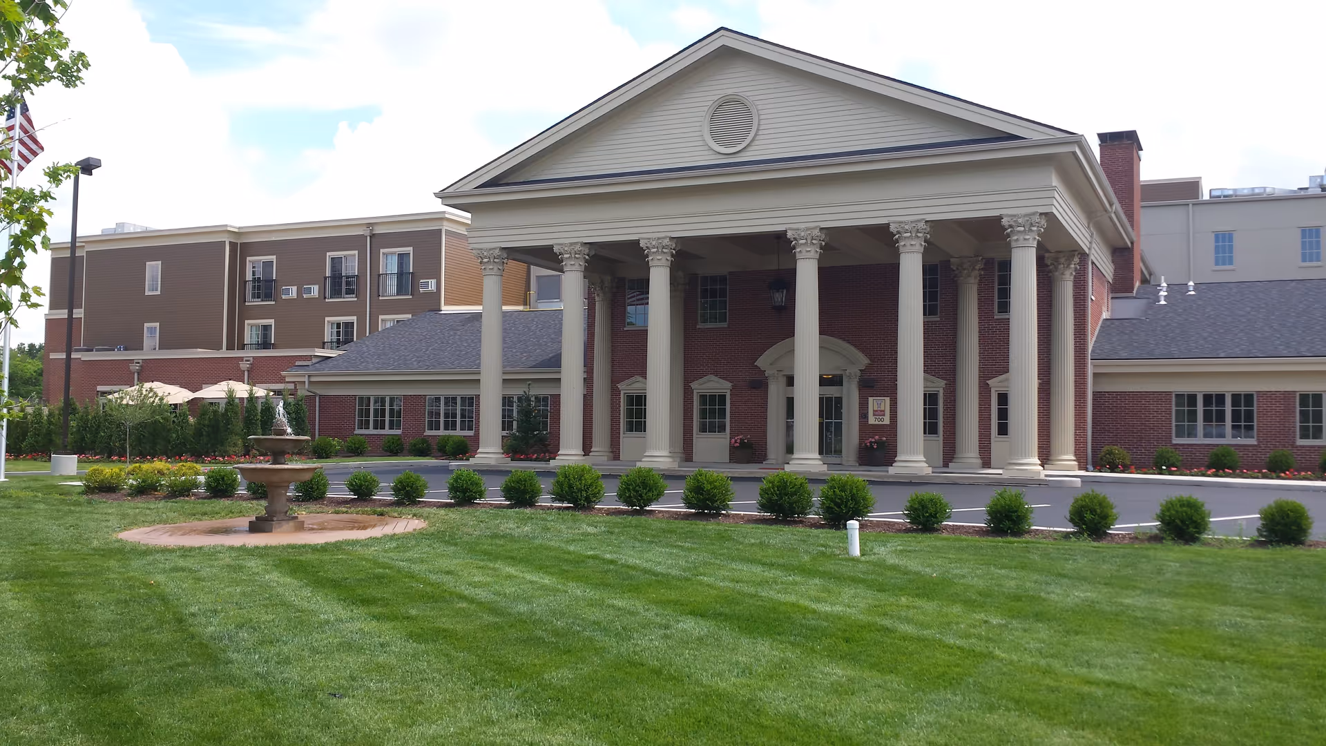 Front exterior view of The Colonnade Senior Living building featuring a large entrance with tall white columns, a manicured lawn with a small fountain, and an American flag on a pole to the left.