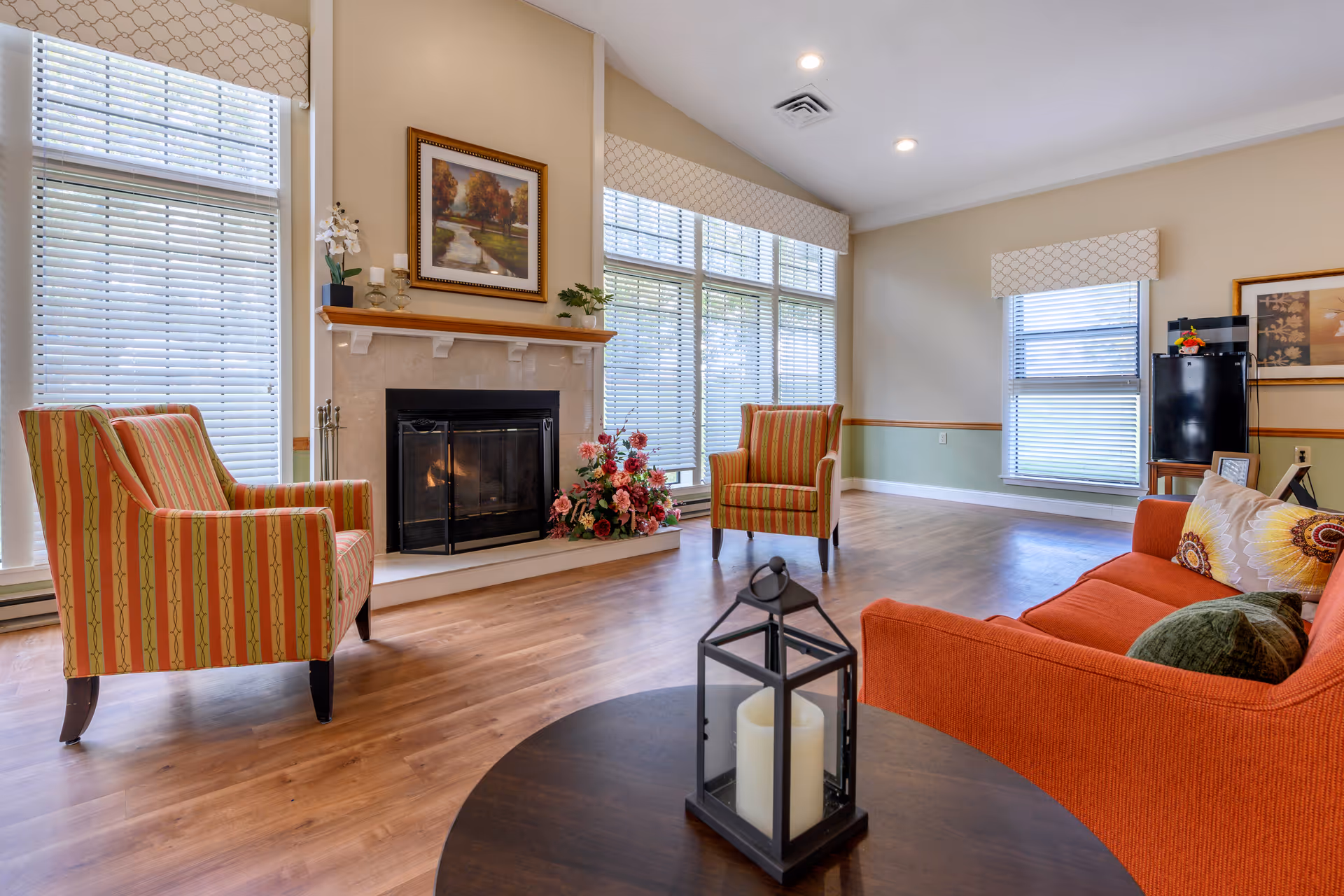 A bright and spacious living room with large windows covered by white blinds, two striped armchairs facing a fireplace with a painting above it, an orange sofa with decorative pillows, and a round wooden table with a black lantern candle holder in the foreground. The room has light-colored walls, wood flooring, and recessed ceiling lights.