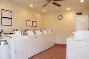 Laundry room with multiple white washing machines and dryers lined up against the walls, beige walls with framed instructions, a ceiling fan, and a clock on the wall.