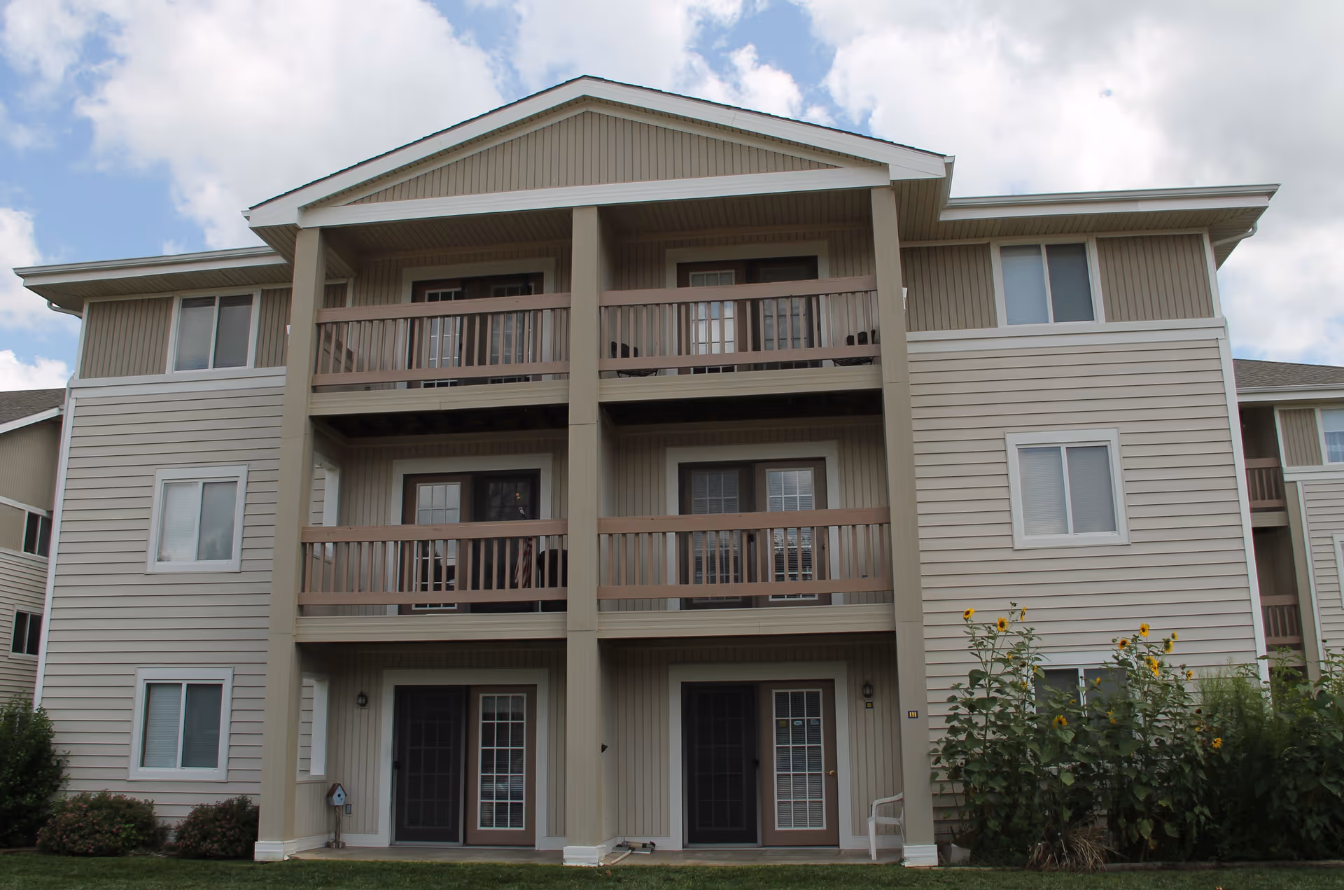 Front exterior of a three-story beige apartment building with stacked balconies, ground-level doors, and sunflowers by the entrance.