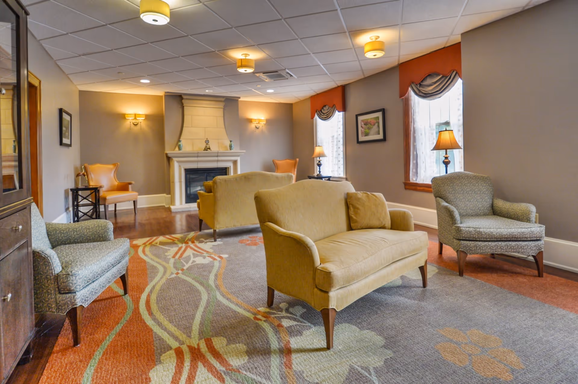 A cozy living room with a fireplace, featuring two beige loveseats, two patterned armchairs, a colorful area rug with floral and wavy designs, wall sconces, table lamps, and windows with decorative valances.