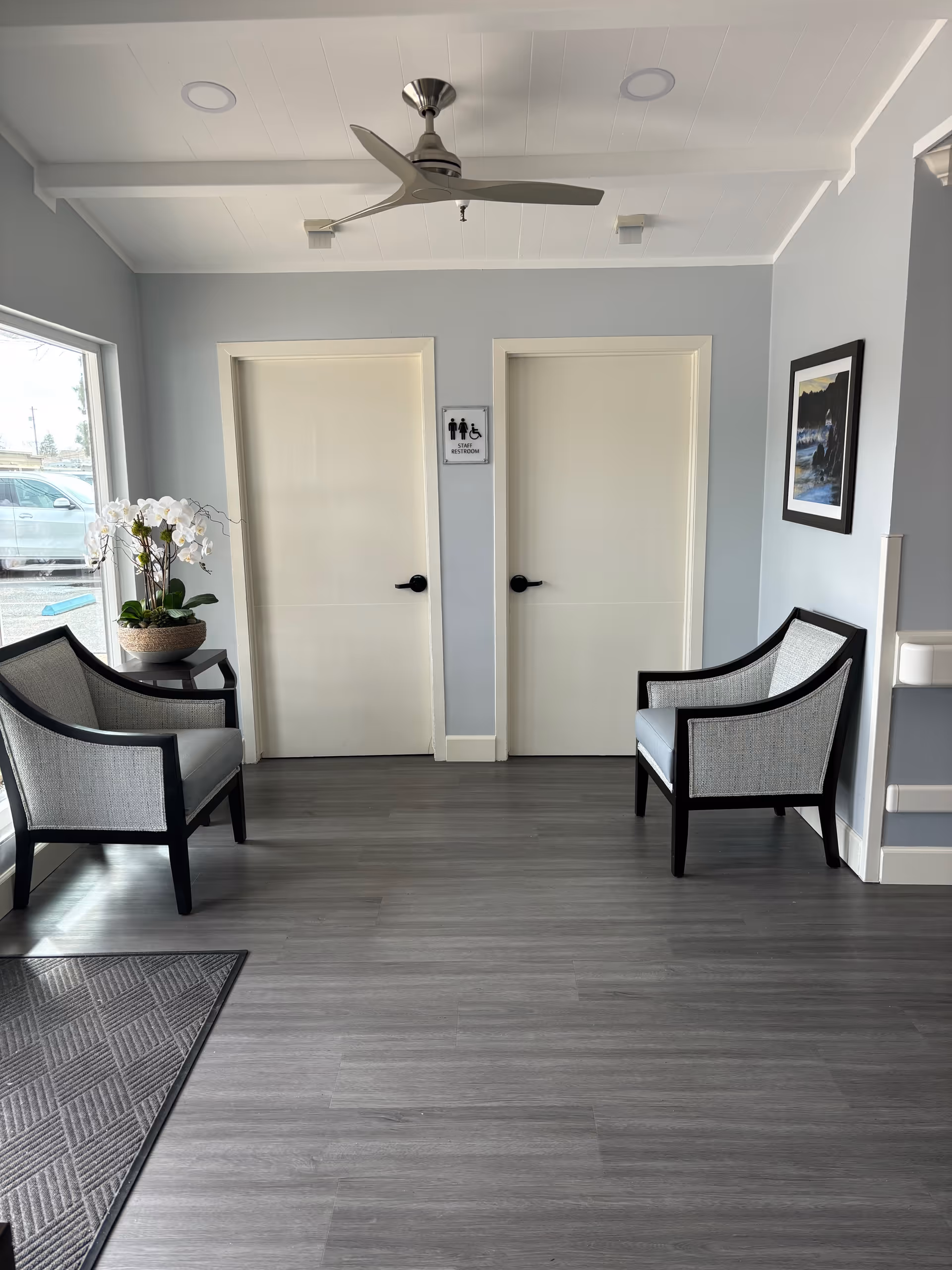 A small waiting area with two gray cushioned chairs facing each other, a small table with a potted orchid plant, two closed white doors with black handles, a sign indicating a restroom for men, women, and wheelchair users, a ceiling fan, and a framed picture on the wall. The floor is gray wood, and there is a large window showing a parked car outside.