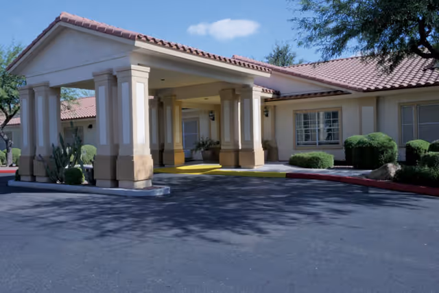 Exterior view of Brookdale Central Chandler facility showing a covered entrance with columns, beige walls, red tile roof, and surrounding landscaping including bushes and a cactus under a blue sky.