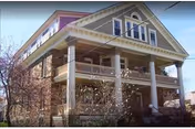 Front exterior of a large historic multi-story brick house with wraparound porches, white columns, and a gabled roof.