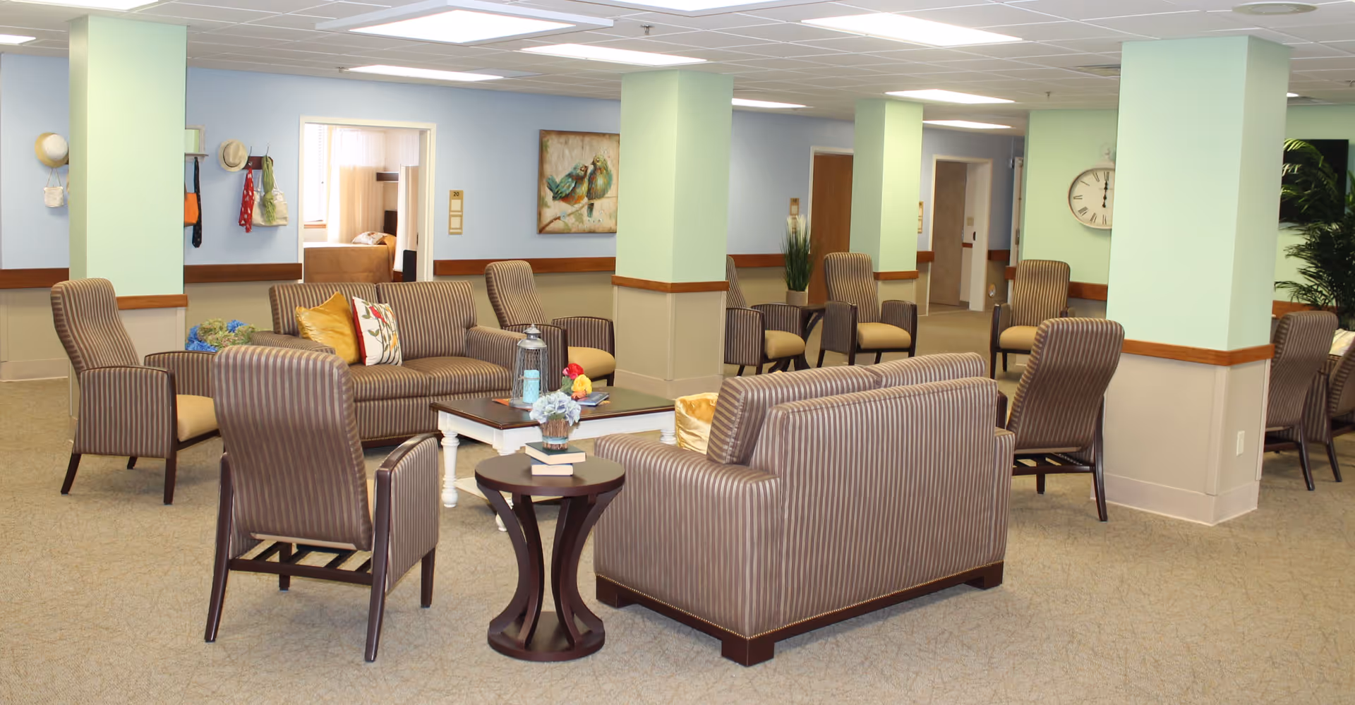 A spacious senior living facility common area with multiple striped armchairs and sofas arranged around coffee tables. The room has light green and beige walls with wooden trim, a large clock on one wall, and decorative plants and artwork. The area is well-lit with ceiling lights and has doorways leading to other rooms.