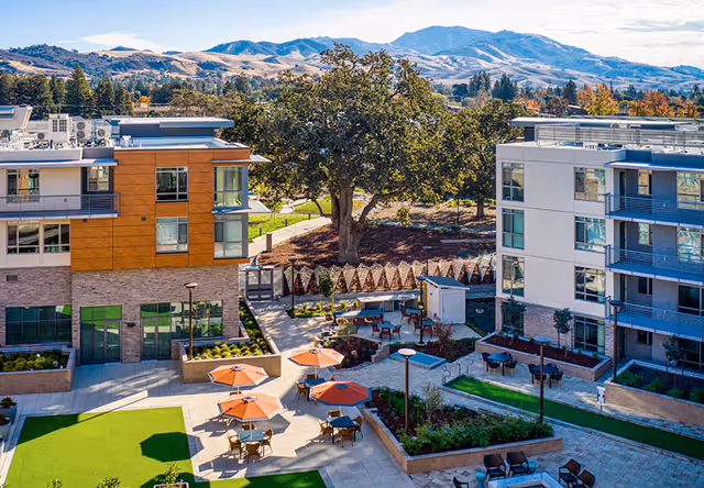 Outdoor courtyard area of Sequoia Living Viamonte at Walnut Creek featuring seating with orange umbrellas, tables, landscaped garden beds, and a large tree in the center, surrounded by modern multi-story residential buildings with mountains visible in the background under a partly cloudy sky.