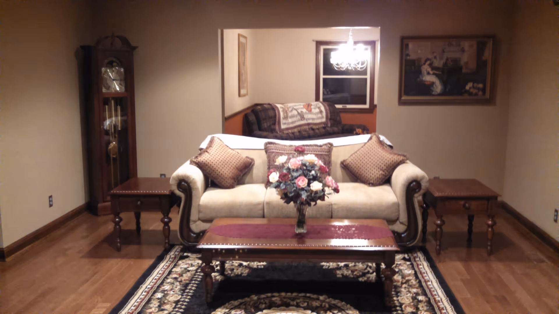 A cozy living room with a beige sofa adorned with three decorative pillows and a floral centerpiece on a wooden coffee table. The room features wooden side tables on either side of the sofa, a grandfather clock in the corner, a patterned rug on the wooden floor, and a painting on the wall. In the background, there is another seating area with a plaid couch and a chandelier hanging from the ceiling.