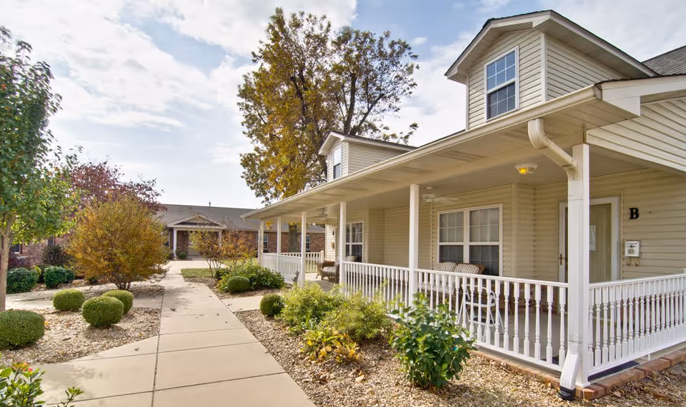 Exterior view of a senior living facility with a beige two-story building featuring a covered porch with white railings and outdoor seating. The walkway is surrounded by landscaped bushes and trees under a partly cloudy sky.