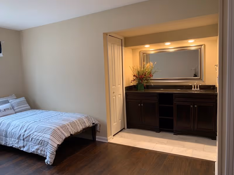A bedroom with a bed on the left side featuring striped bedding and a pillow with the word 'home'. On the right side, there is a built-in vanity area with a large mirror, dark wood cabinets, a sink, and a floral arrangement. The floor is dark wood with a tiled section in front of the vanity.