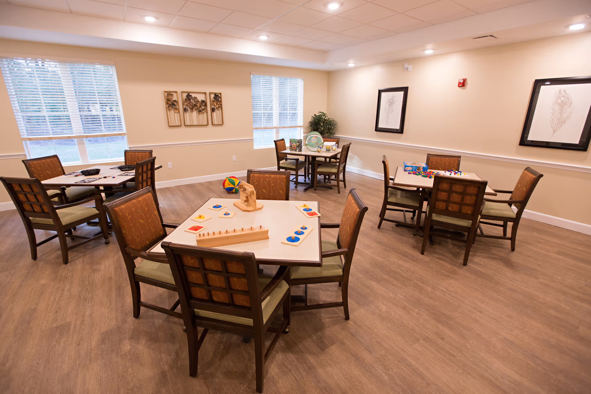 A well-lit activity room with several square tables and chairs arranged around them. Each table has various games and puzzles on top. The room has large windows with blinds, light-colored walls adorned with framed artwork, and a wooden floor.