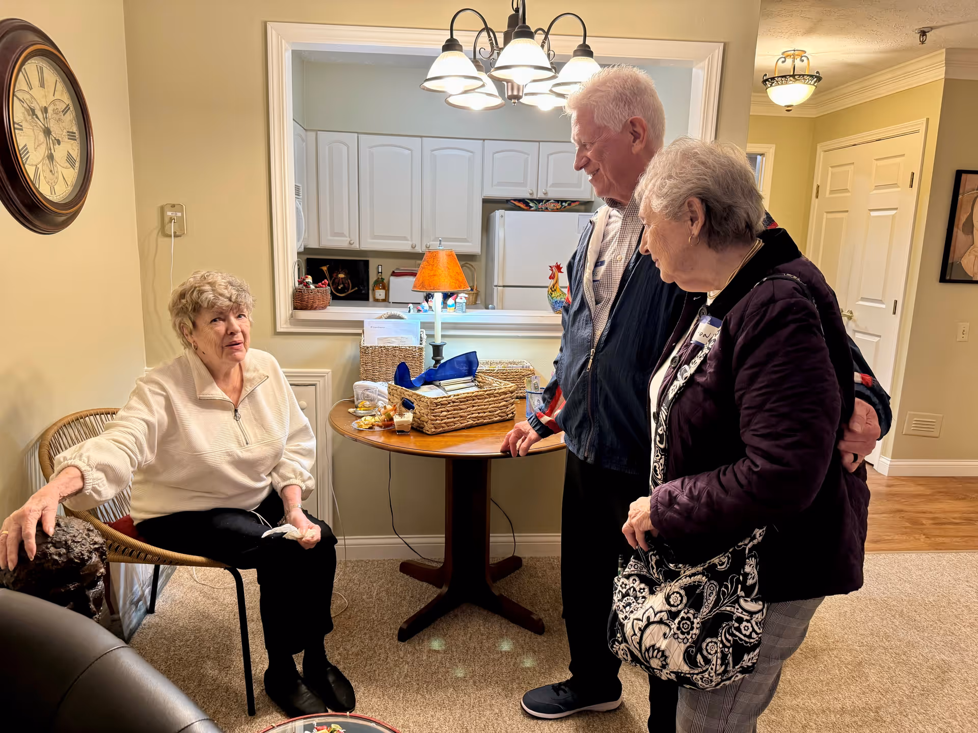 An elderly woman sitting on a wicker chair in a cozy room, talking to an elderly couple standing nearby. Behind them is a small round table with baskets and a lamp, and a kitchen area with white cabinets and a refrigerator is visible through a pass-through window.