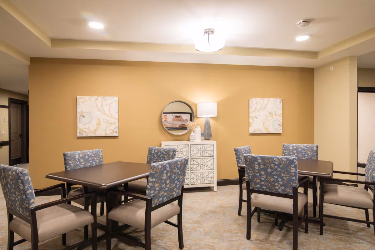 A cozy interior room with two square wooden tables, each surrounded by four cushioned chairs with blue floral upholstery. The walls are painted a warm beige color and decorated with two floral-patterned square paintings and a round mirror above a white decorative cabinet. A table lamp with a white shade sits on the cabinet, providing soft lighting.