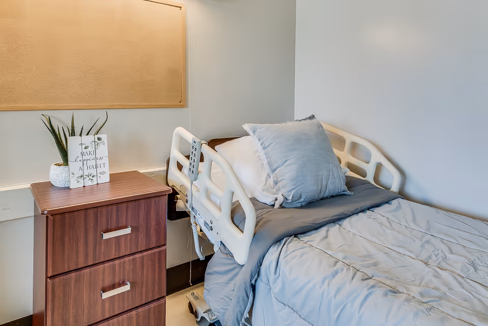 A medical-style bed with pillows and gray bedding beside a wooden bedside cabinet topped with a plant and decorative sign.