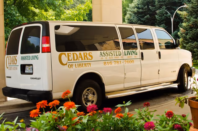 White 'Cedars of Liberty' assisted living passenger van parked at a facility entrance with colorful flowers in the foreground.