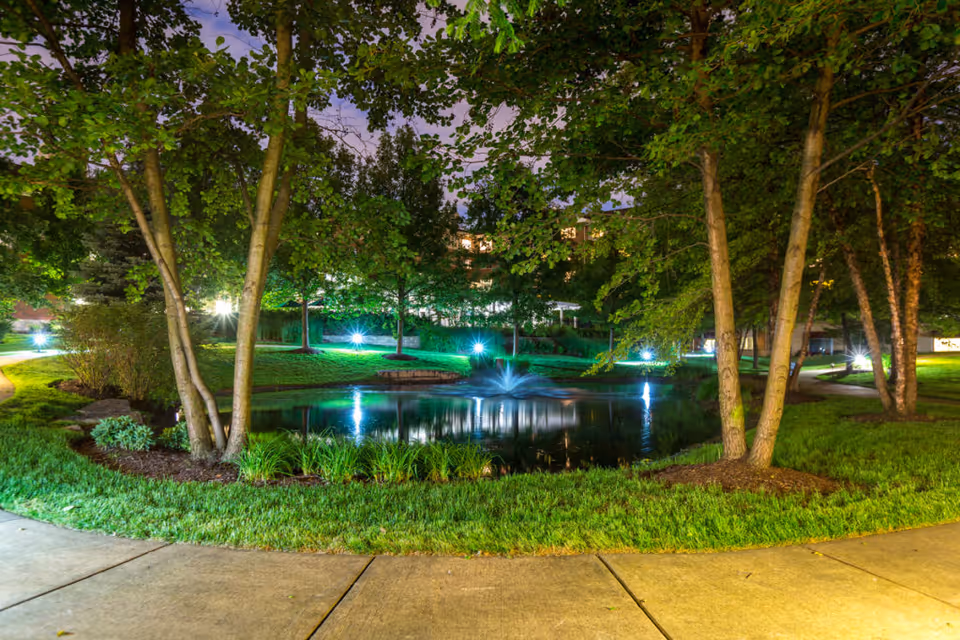 A nighttime landscaped courtyard with trees, an illuminated pond and fountain surrounded by grass and paved walkways.
