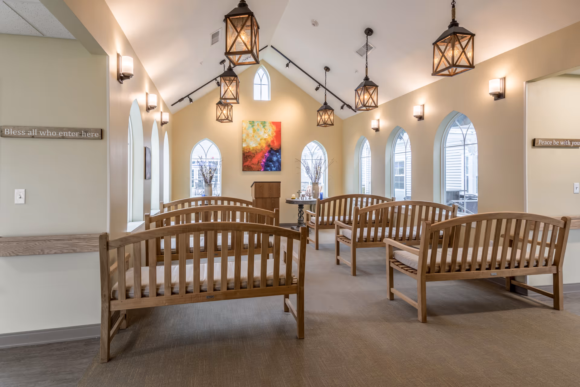 A bright and airy room with high vaulted ceilings and arched windows. The room contains several wooden benches with cushions arranged in rows facing a small podium and a colorful abstract painting on the far wall. Lantern-style pendant lights hang from the ceiling, and wall sconces provide additional lighting. Signs on the walls read 'Bless all who enter here' and 'Peace be with you'.