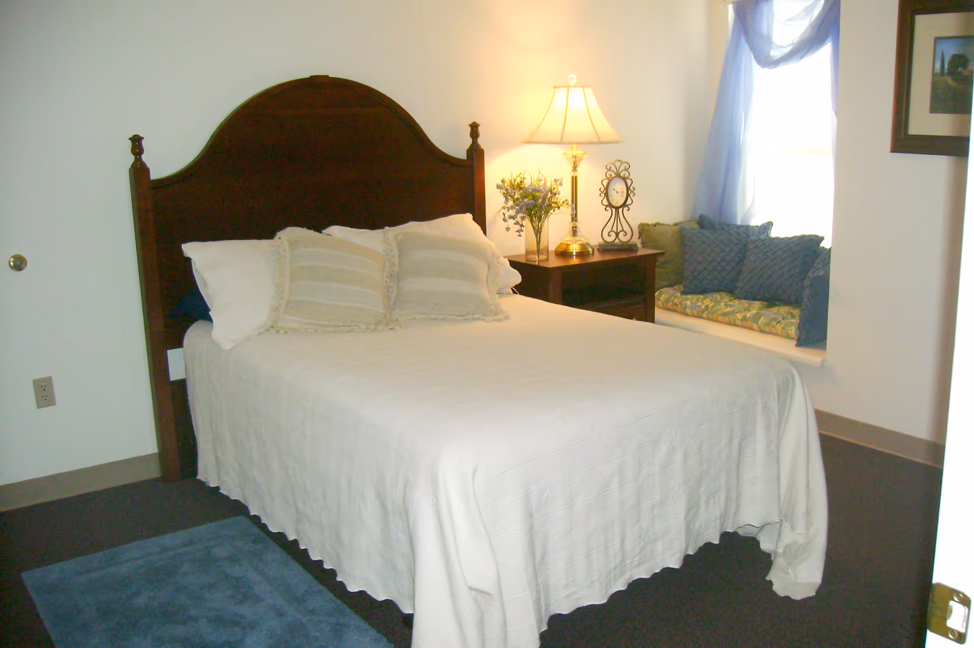 A bedroom with a neatly made bed featuring a wooden headboard, white bedspread, and three pillows. Next to the bed is a wooden nightstand with a lamp, a vase of flowers, and a decorative clock. A window seat with cushions is visible by the window with sheer blue curtains.
