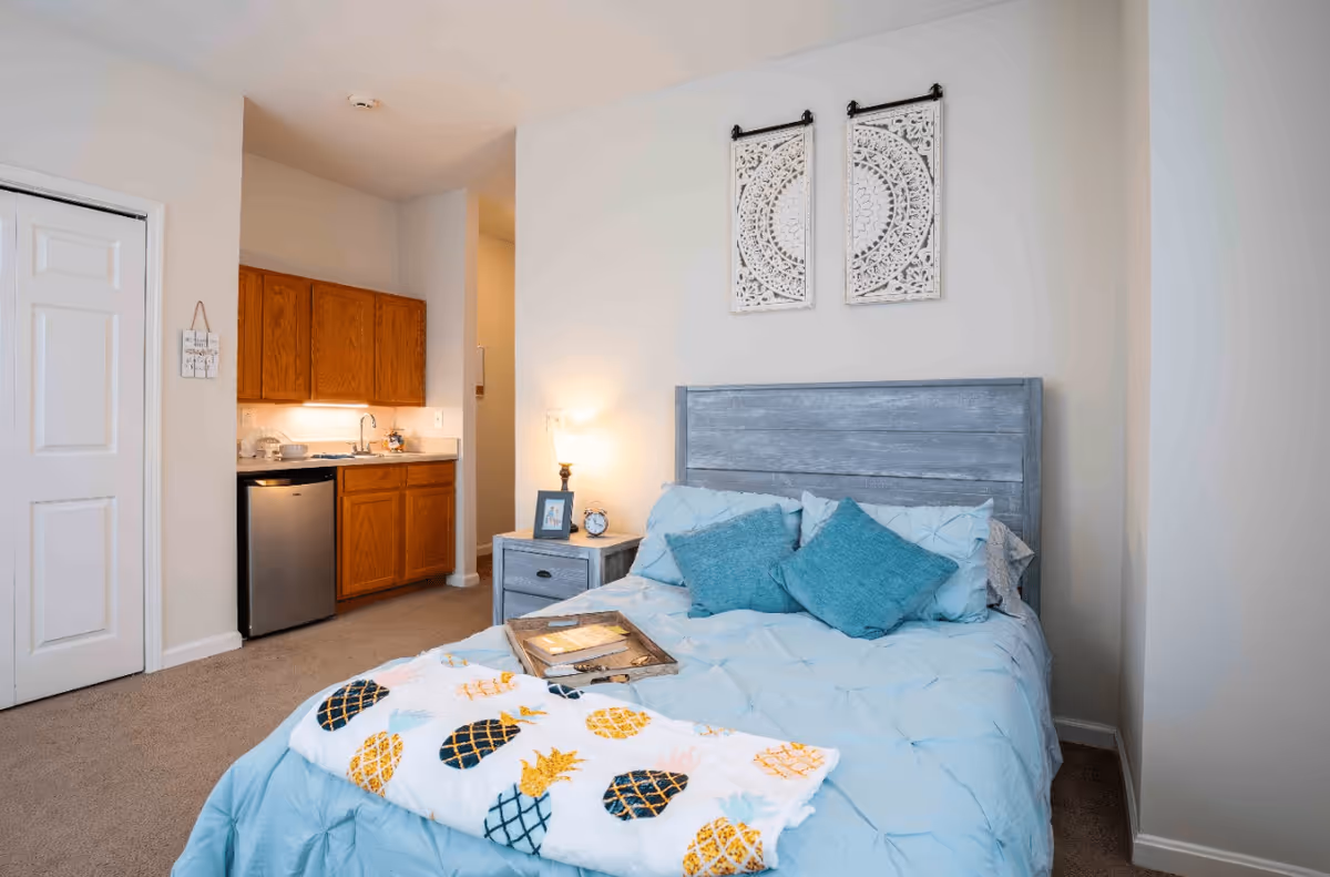 A cozy bedroom with a blue bedspread and two teal pillows on a wooden bed frame. A colorful pineapple-patterned blanket is draped at the foot of the bed. Next to the bed is a small nightstand with a lamp, a framed photo, and an alarm clock. In the background, there is a small kitchenette with wooden cabinets, a sink, and a mini refrigerator. The walls are light-colored and two decorative white wall hangings are above the bed.