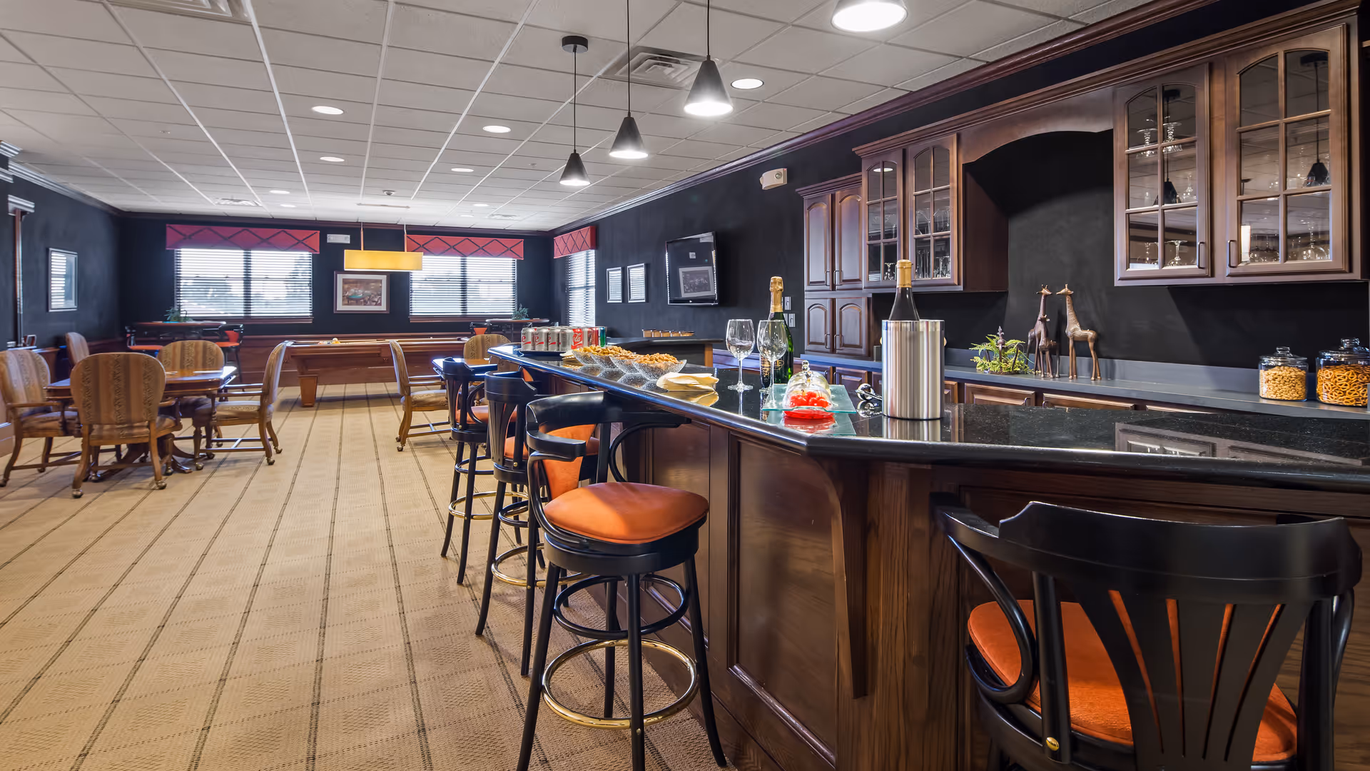 A spacious room with a long wooden bar counter with black granite top and several bar stools with orange cushions. Behind the bar are wooden cabinets with glass doors displaying glassware. On the counter are bottles of wine, glasses, snacks, and a wine cooler. In the background, there are round tables with chairs and a pool table near the windows with red valances. The room has a carpeted floor and pendant lights hanging from the ceiling.