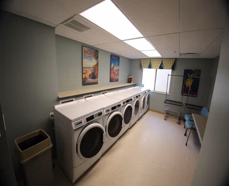 A laundry room with six front-loading washing machines lined up against a wall. The room has light green walls, a window with a valance, a trash bin, two chairs, a folding table, and a laundry cart. There are three colorful posters on the walls.