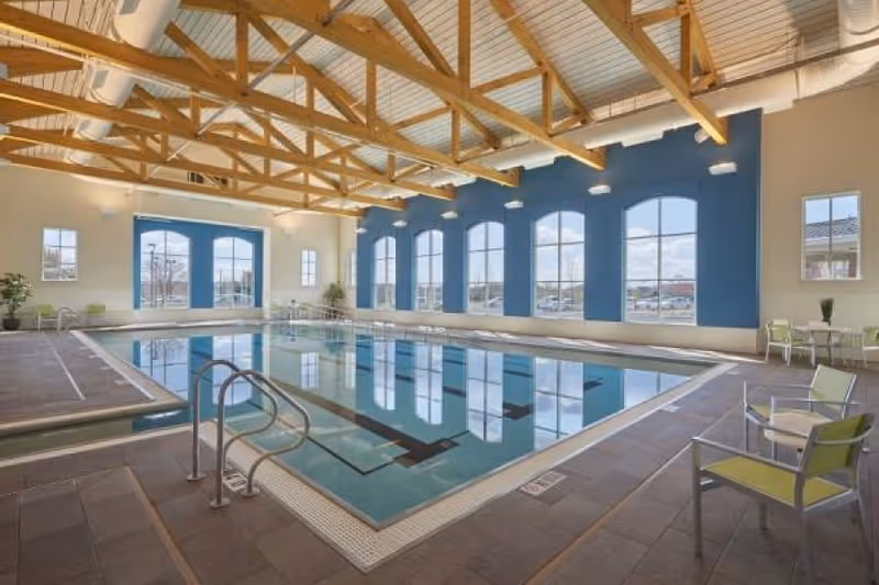 Indoor swimming pool in a bright room with arched windows, exposed wooden ceiling beams, and seating along the sides.