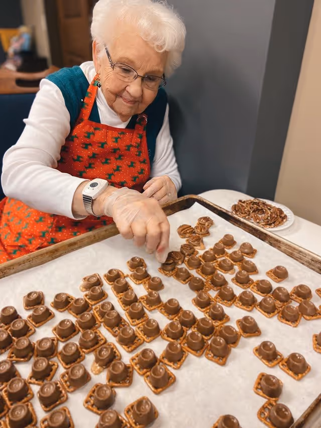 An elderly woman wearing glasses, a white long-sleeve shirt, and a red apron with green patterns is placing chocolate candies on pretzel squares arranged on a baking tray lined with parchment paper. A plate of pecans is visible on the table beside her.