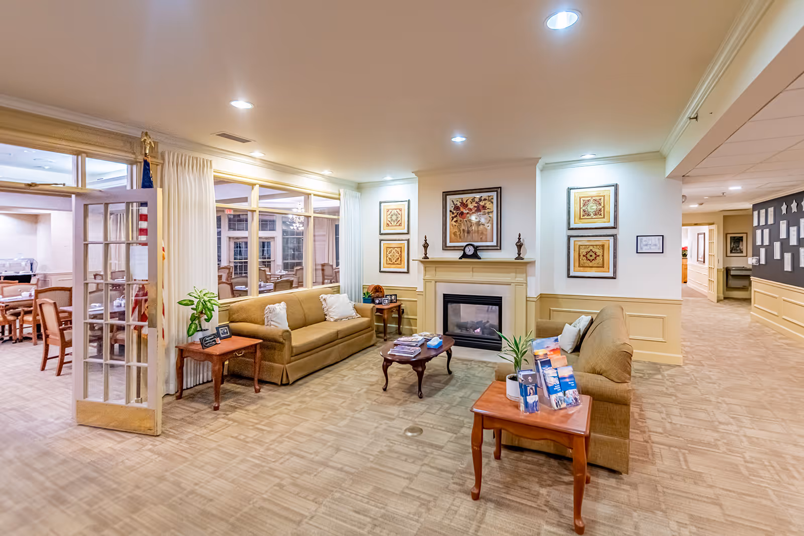 A cozy living room area in a senior living facility with two brown sofas facing each other, a wooden coffee table in the center, and a fireplace with framed artwork above it. There are side tables with plants and brochures, and large windows with white curtains looking into a dining room with tables and chairs.