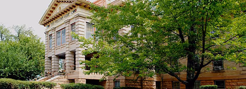 Exterior view of a large, historic stone building partially obscured by green trees and bushes. The building has multiple windows and a prominent entrance with columns and a triangular pediment. The name 'Friendship House' is visible on the building's facade.