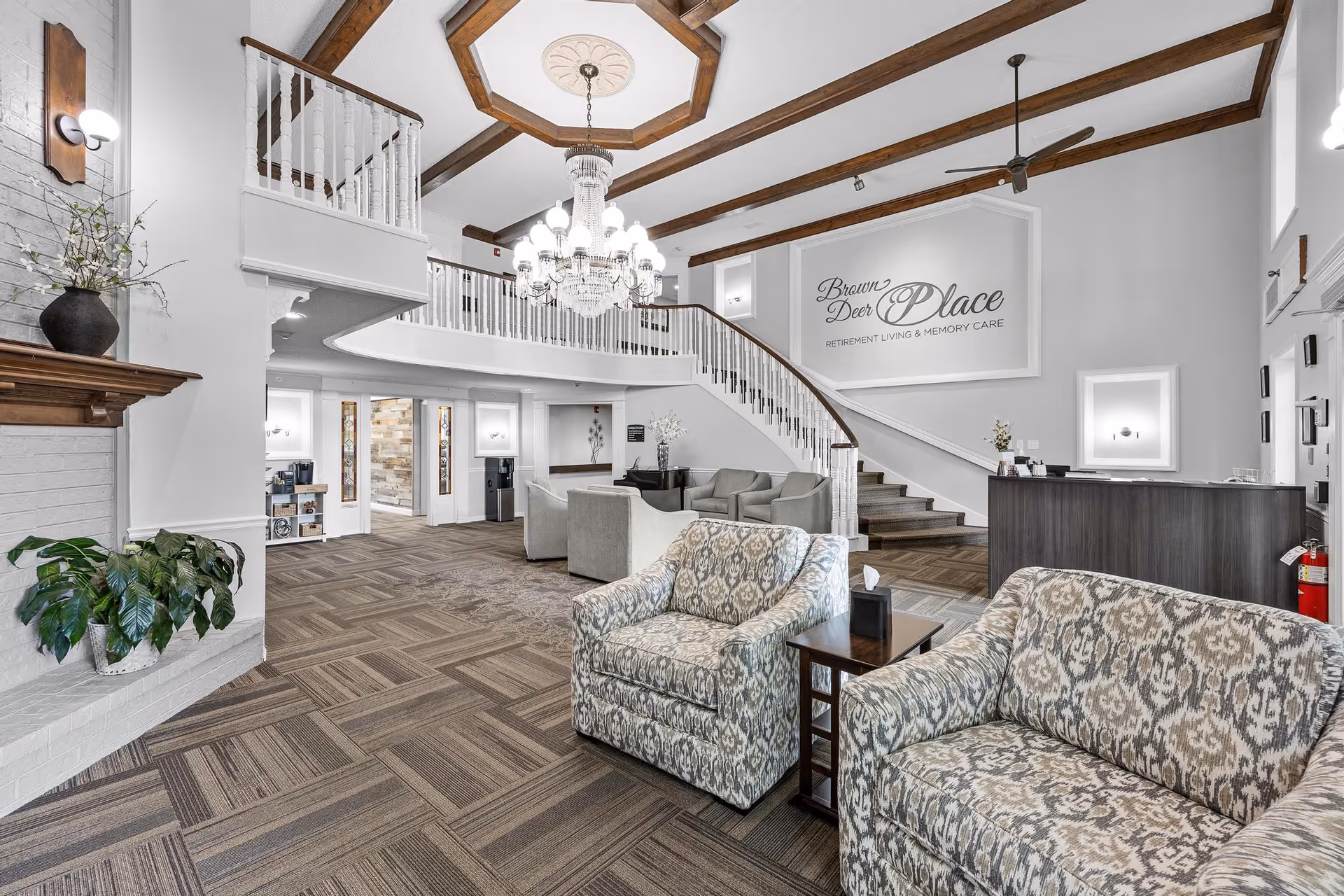 Spacious and well-lit senior living facility lobby with patterned armchairs and sofas arranged around small tables, a large chandelier hanging from a ceiling with wooden beams, a curved staircase with white railings, and a sign on the wall that reads 'Brown Deer Place Retirement Living & Memory Care'.