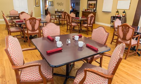 A senior living dining room with multiple tables, pink patterned chairs, and napkin settings.