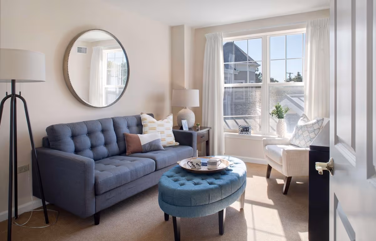 Bright living room with a tufted gray sofa, round mirror, blue ottoman, armchair by a large window and a floor lamp.