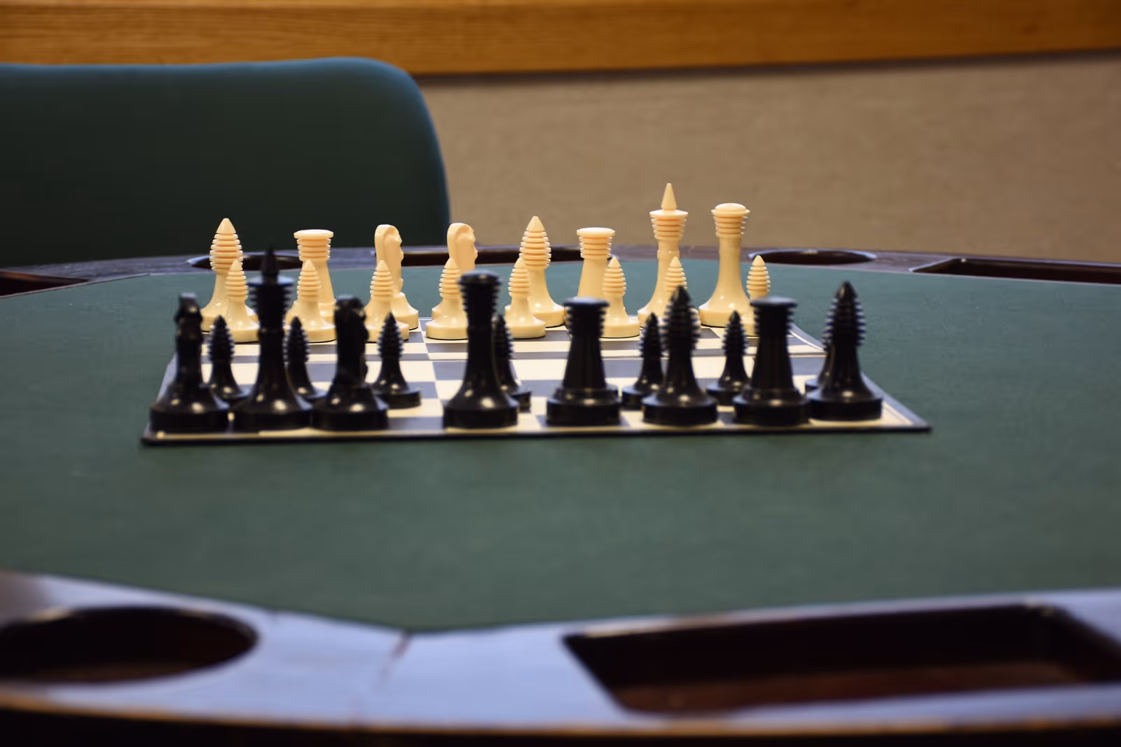 A chessboard set up for a game with black and white chess pieces on a green felt table, with a green chair and a wooden wall in the background.