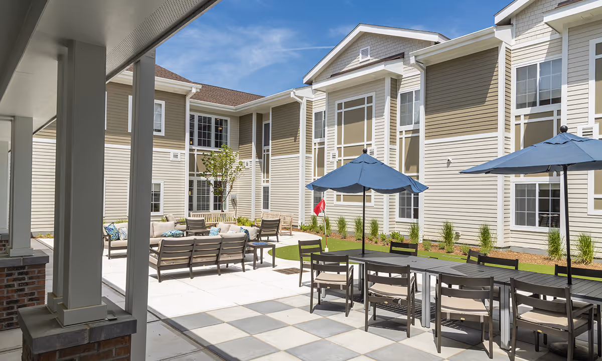 Sunny courtyard with outdoor tables, blue umbrellas, lounge seating, and the beige-sided senior living building in the background.