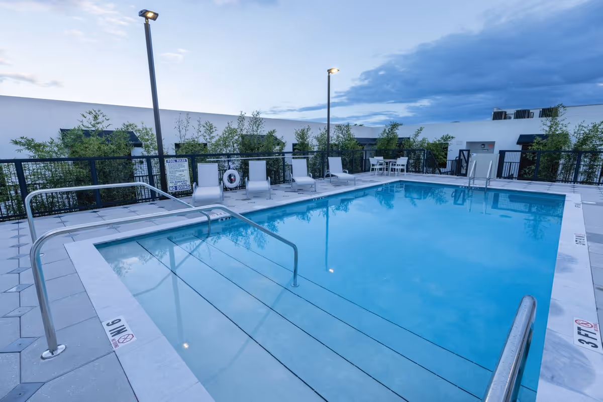 Outdoor swimming pool area at dusk with clear blue water, surrounded by a tiled deck with white lounge chairs and a small table with chairs. The pool has metal handrails and depth markers visible. There are tall plants and a white building wall in the background under a partly cloudy sky.