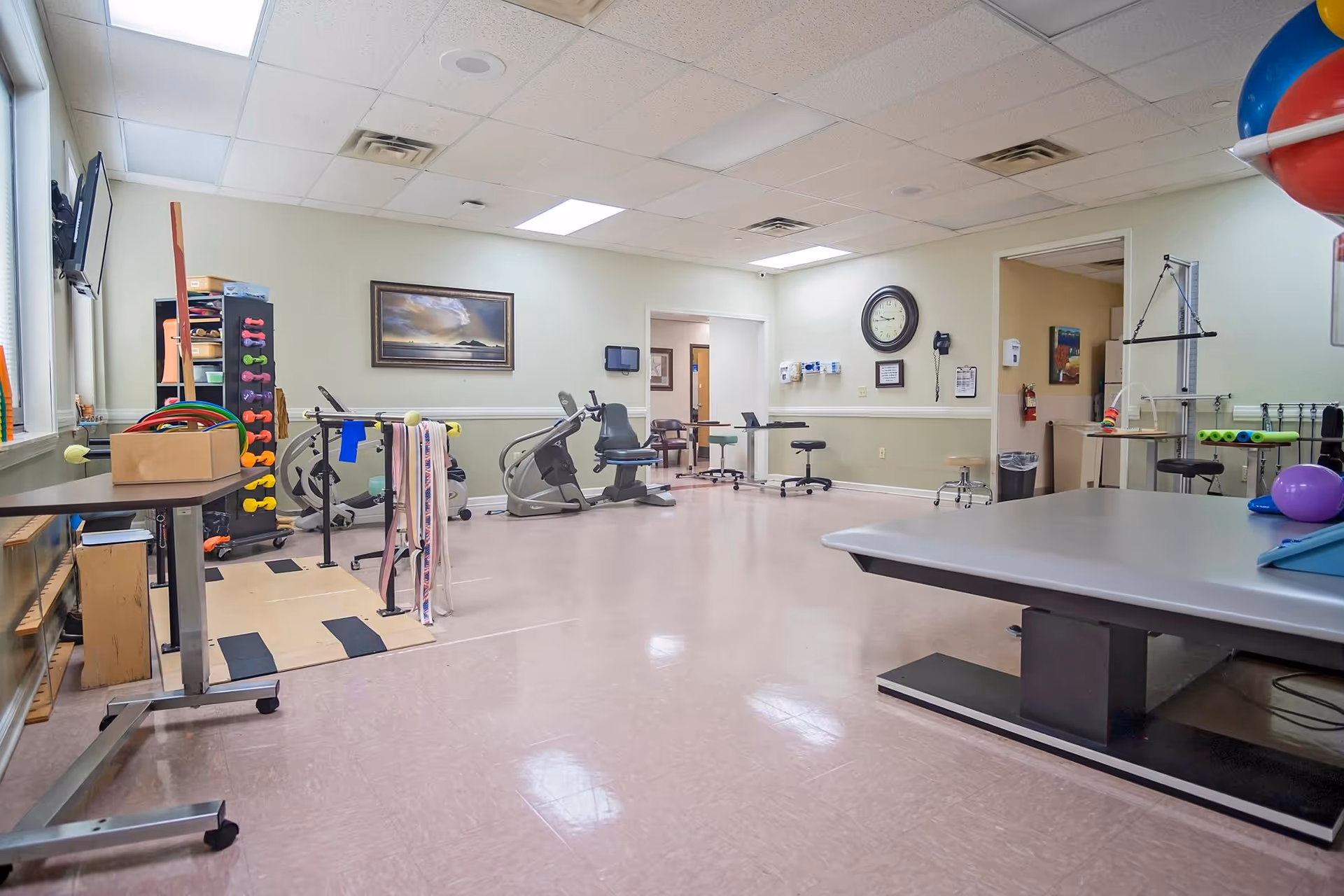 A spacious therapy or rehabilitation room with exercise equipment including stationary bikes, weights, therapy tables, and various physical therapy tools. The room has a tiled floor, light green walls, a clock on the wall, and fluorescent ceiling lights. There are chairs and small tables along the walls, and a framed picture hangs on one wall.