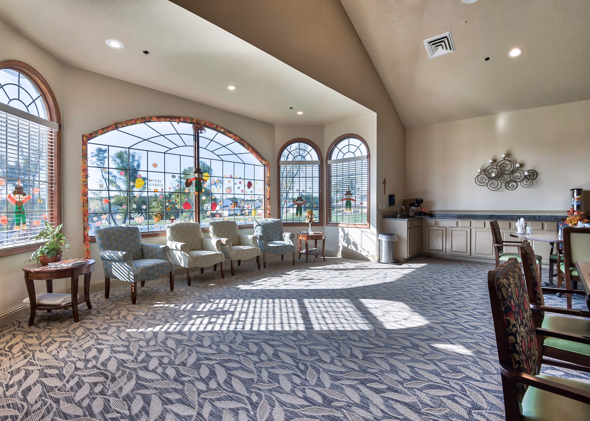 A bright, spacious common area in Oak Pointe Assisted Living with large arched windows decorated with autumn-themed stickers. The room features several upholstered armchairs arranged near the windows, a small wooden side table with a plant, and a kitchenette area with cabinets, a coffee maker, and a water dispenser. The carpet has a leaf pattern, and sunlight streams through the windows, casting shadows on the floor.