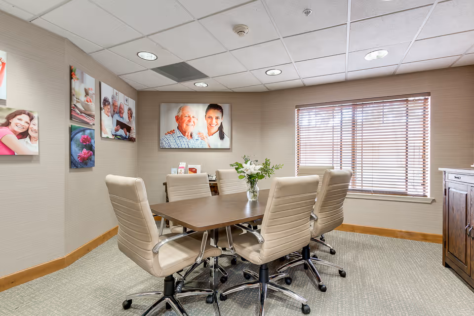 Small conference room with a rectangular table surrounded by beige rolling chairs, wall photos, and a window with blinds.
