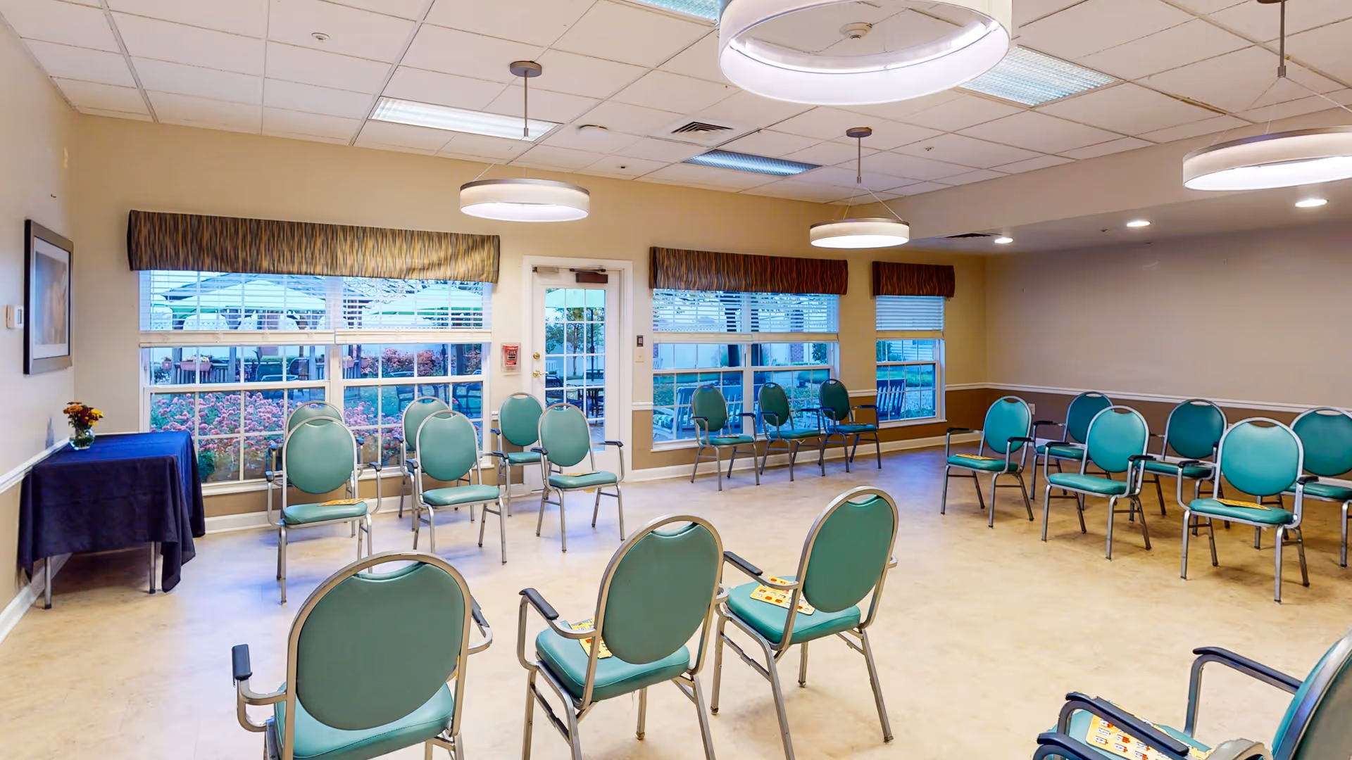 A spacious room with multiple green chairs arranged in a semi-circle facing a table covered with a dark blue cloth. Large windows with brown valances allow natural light to enter, showing an outdoor area with umbrellas and greenery. The ceiling has several round hanging lights and fluorescent panels.