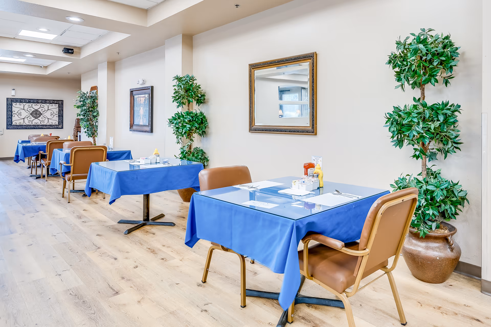 Dining room with several tables covered in blue tablecloths, brown chairs, potted plants, and wall decor in a bright interior.
